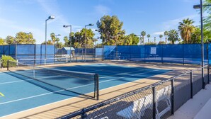 Un court de tennis en plein air, entouré d’une clôture en treillis métallique, d’arbres et de lampadaires.