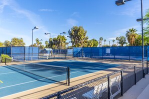 An outdoor tennis court with a chain-link fence, surrounded by trees and street lamps.
