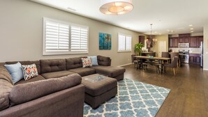 A spacious living room with a sectional sofa, a dining area, and a kitchen in the background. - Family Oasis on 1/4 acre! Pool, putting green + swingset! 30 min to Joshua Tree! (Indio)