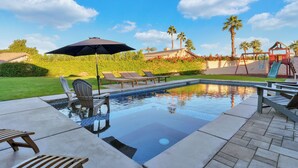 A swimming pool with lounge chairs and an umbrella, surrounded by palm trees and a house. - Family Oasis on 1/4 acre! Pool, putting green + swingset! 30 min to Joshua Tree! (Indio)