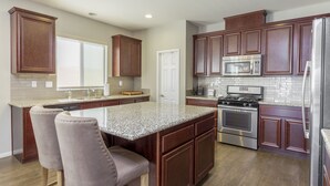 A kitchen with dark wooden cabinets, a granite countertop, stainless steel appliances, and two bar stools. - Family Oasis on 1/4 acre! Pool, putting green + swingset! 30 min to Joshua Tree! (Indio)