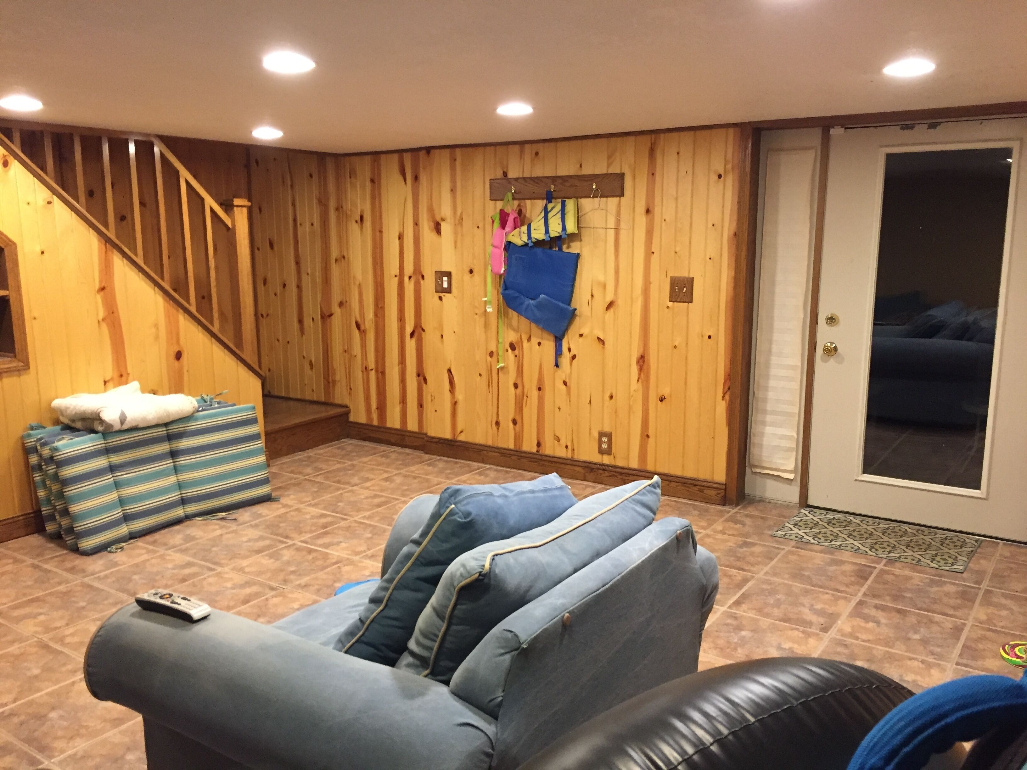 A living room with a wooden wall, a sofa, a striped ottoman, and a remote control on the sofa.