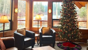 A spacious room with a high ceiling, large windows, and a Christmas tree. There are two armchairs and a sofa. - Heaven Can Wait Lodge ~ Riverfront Estate on 12 Private Acres ~ Expansive Views (Leavenworth)