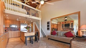 Une cuisine spacieuse avec un îlot central, un coin repas avec des tabourets, un salon avec un canapé et des coussins, et un balcon avec une balustrade.