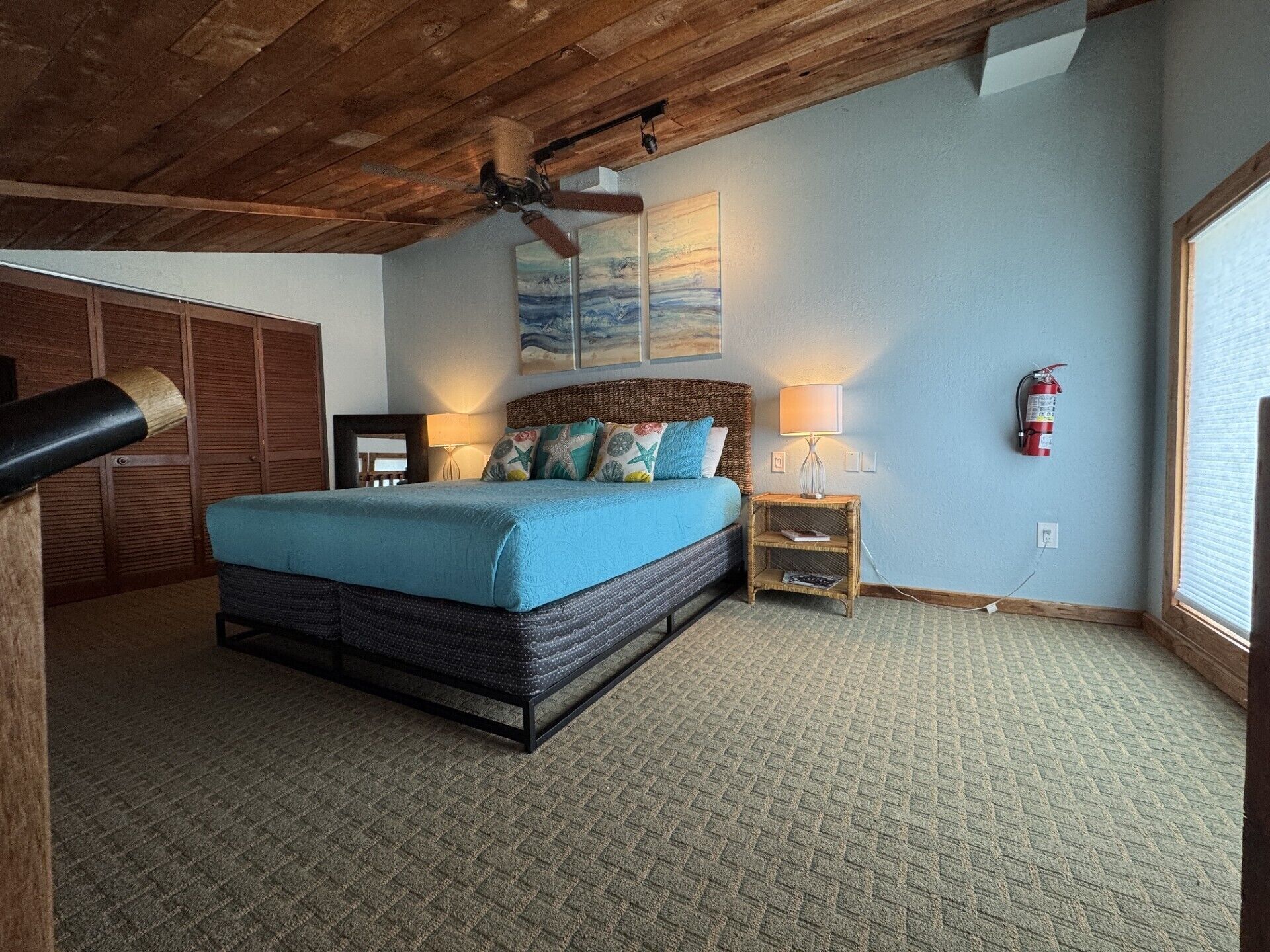 Une chambre à coucher comprenant un lit, un ventilateur de plafond, une table de chevet avec une lampe, un extincteur et une fenêtre.