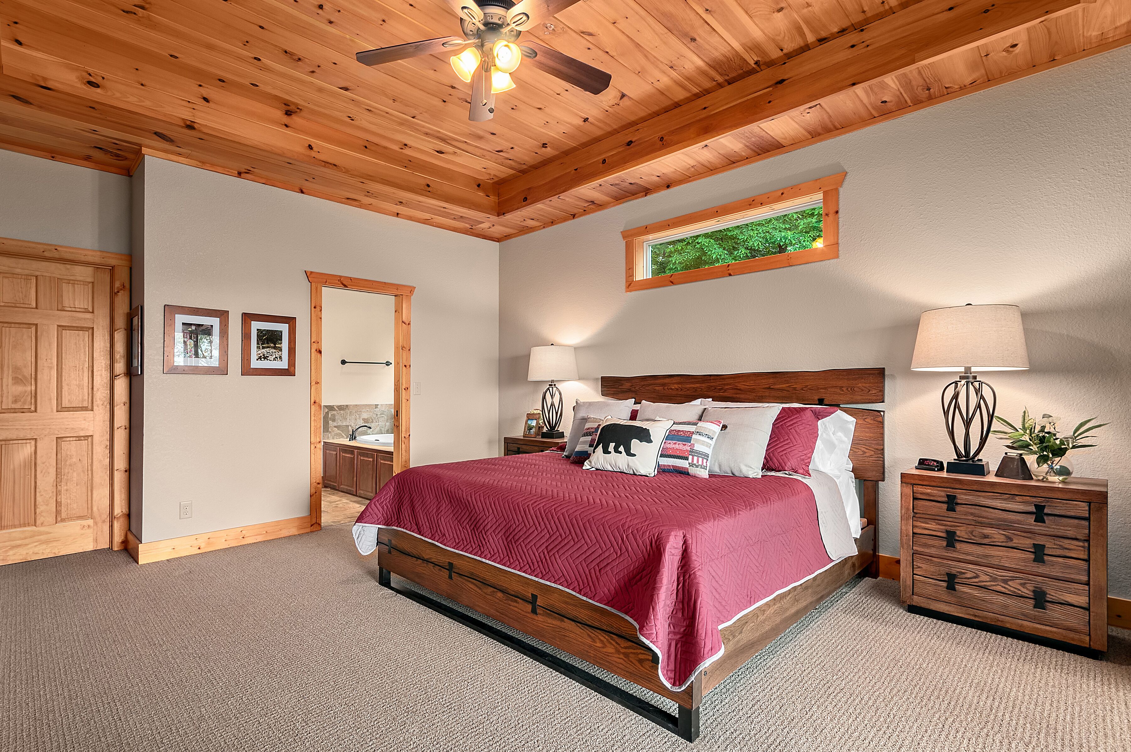 A bedroom with a wooden ceiling, a bed with a red bedspread, a nightstand with a lamp, and a dresser.