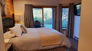 Une chambre avec un grand lit, une tête de lit en bois, un téléviseur fixé au mur et une vue sur l’extérieur par les fenêtres.