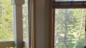Une pièce dotée d’une grande fenêtre donnant sur une forêt et un étang, une terrasse en bois à l’extérieur et un fauteuil en osier près de la fenêtre.