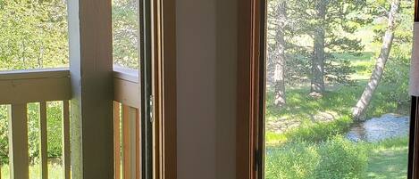 A room with a large window offering a view of a forest and a pond, a wooden deck outside, and a wicker chair near the window.