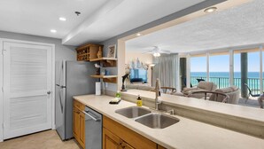 A kitchen with a large window, a refrigerator, a sink, and a dining area with a view of the ocean. - Oceanfront 2BR Condo at Long Beach Resort, Panama City Beach, sleeps 6. (Panama City Beach)