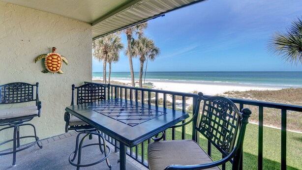 A covered outdoor seating area with a table and chairs, overlooking a beach with palm trees and the ocean. - BEAUTIFUL CONDO WITH PERFECT VIEW OF BEACH AND GULF WITH NEW HEATED POOL (Indian Rocks Beach)