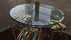 A round glass table with metal chairs, positioned in a corner with white blinds. - DOWNTOWN COZY APARTMENT (St Augustine)