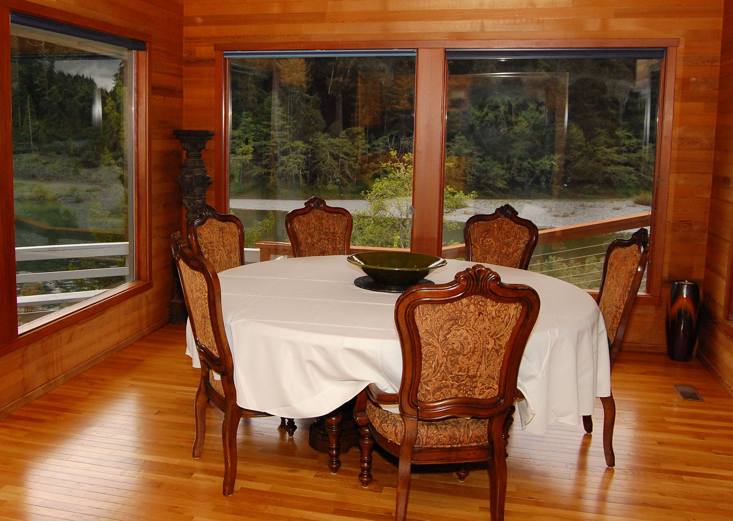 Une salle à manger avec une table ronde, six chaises et une grande fenêtre donnant sur une zone boisée.