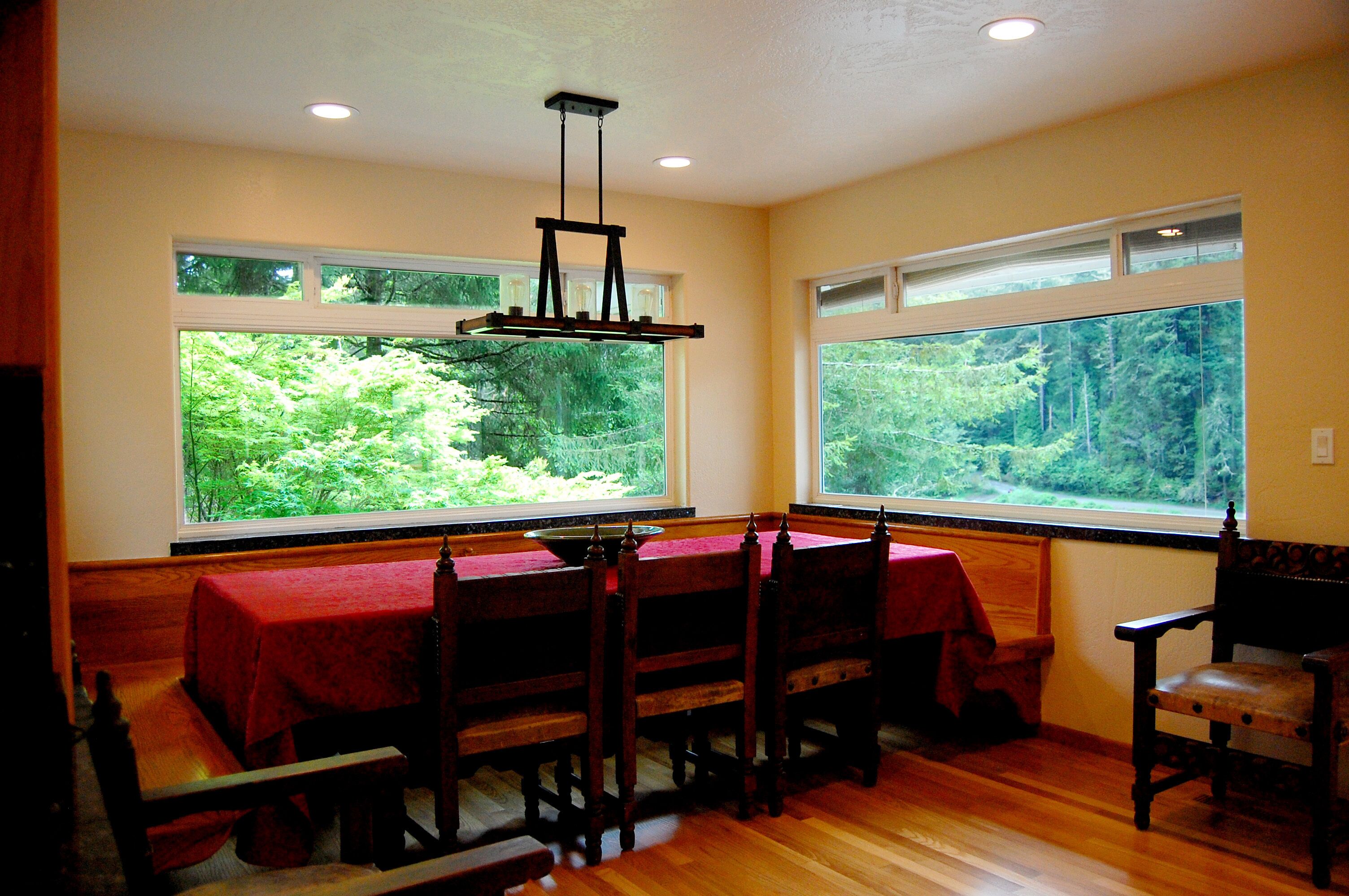 A dining room with a wooden table and chairs, large windows with a view of greenery, and a hanging light fixture.