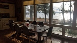 A dining area with a large table and chairs, a kitchen area with cabinets, and a view of a lake through large windows. - Sun, Sand, Snow, Year Round Recreation, With Private Beach (Roscommon)