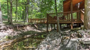 Een houten terras met een eenvoudige blokhut, omgeven door een bebost gebied met een beekje.