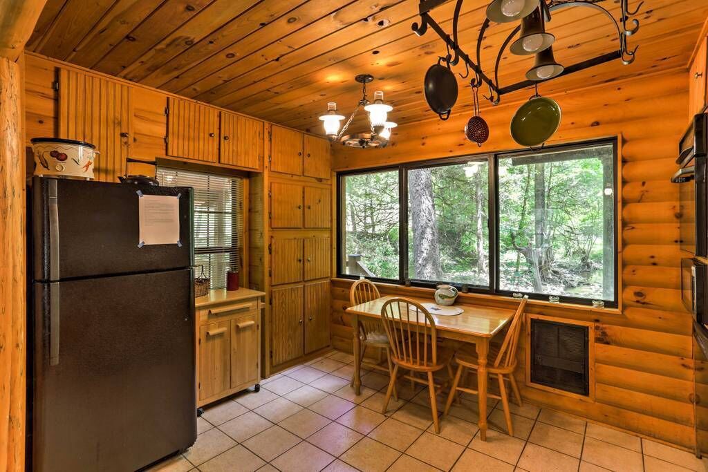 A log cabin kitchen with a black refrigerator, wooden cabinets, a dining table with chairs, and a large window offering a view of the forest.