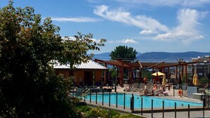 A pool area with lounge chairs and umbrellas, surrounded by trees and a view of mountains. - 5 ⭐️ Unit 437 - Sol Terra Stays - Bright 2 bdrm with Pool & Courtyard Views (Kelowna)
