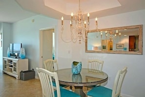 A dining area with a round glass table, white chairs, teal cushions, and a chandelier.