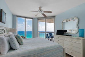 A bedroom with a bed, a desk, a chair, a ceiling fan, a large window with a view of the ocean, and a balcony with a chair and table.