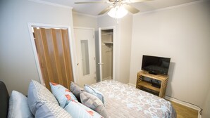 Een slaapkamer met een bed, kussens, een televisie op een houten standaard, een plafondventilator en een kledingkast.