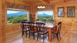 Une salle à manger en bois, avec une table et des chaises, de grandes fenêtres avec des rideaux et des œuvres d’art encadrées accroch ées aux murs.