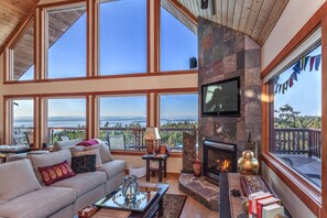 A spacious living room with large windows, a fireplace, and a television mounted on the wall.