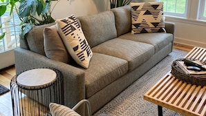 A modern living room with a grey sofa, patterned cushions, a round side table, a coffee table, and large green plants. - Modern Designer Beach House 1 mile to the beach! Pet-friendly & bright (Sawyer)