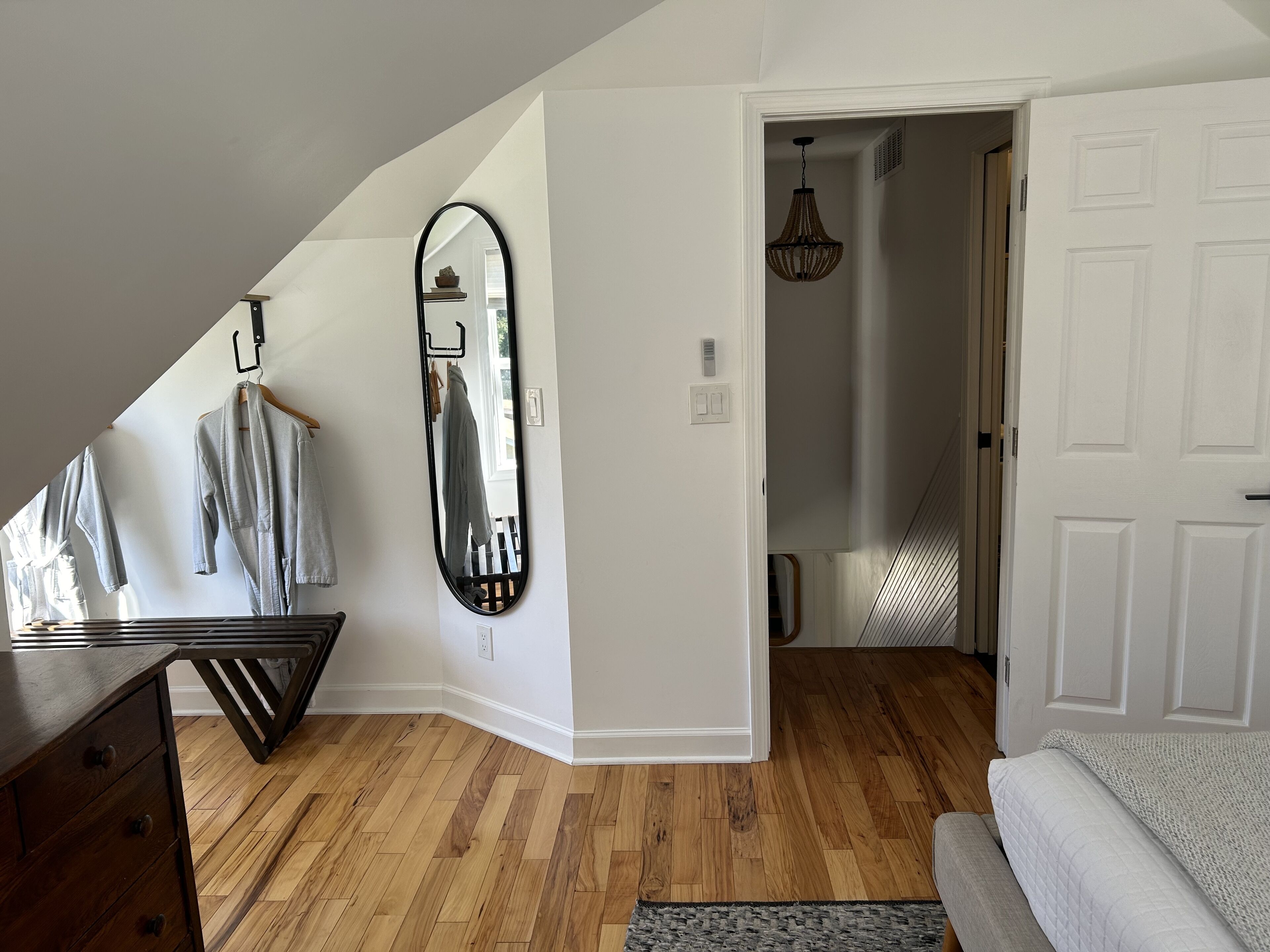 A room with a wooden floor, a large mirror, a hanging light fixture, and a door leading to another room.