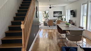 A spacious living area with a wooden staircase, a dining table with chairs, a sofa, and a television mounted on the wall. - Modern Designer Beach House 1 mile to the beach! Pet-friendly & bright (Sawyer)