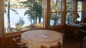 A dining area with a round table covered by a floral tablecloth, surrounded by wooden chairs. Large windows provide a view of a lake and trees.