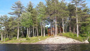 A cabin surrounded by tall pine trees, a sandy beach, and a body of water.