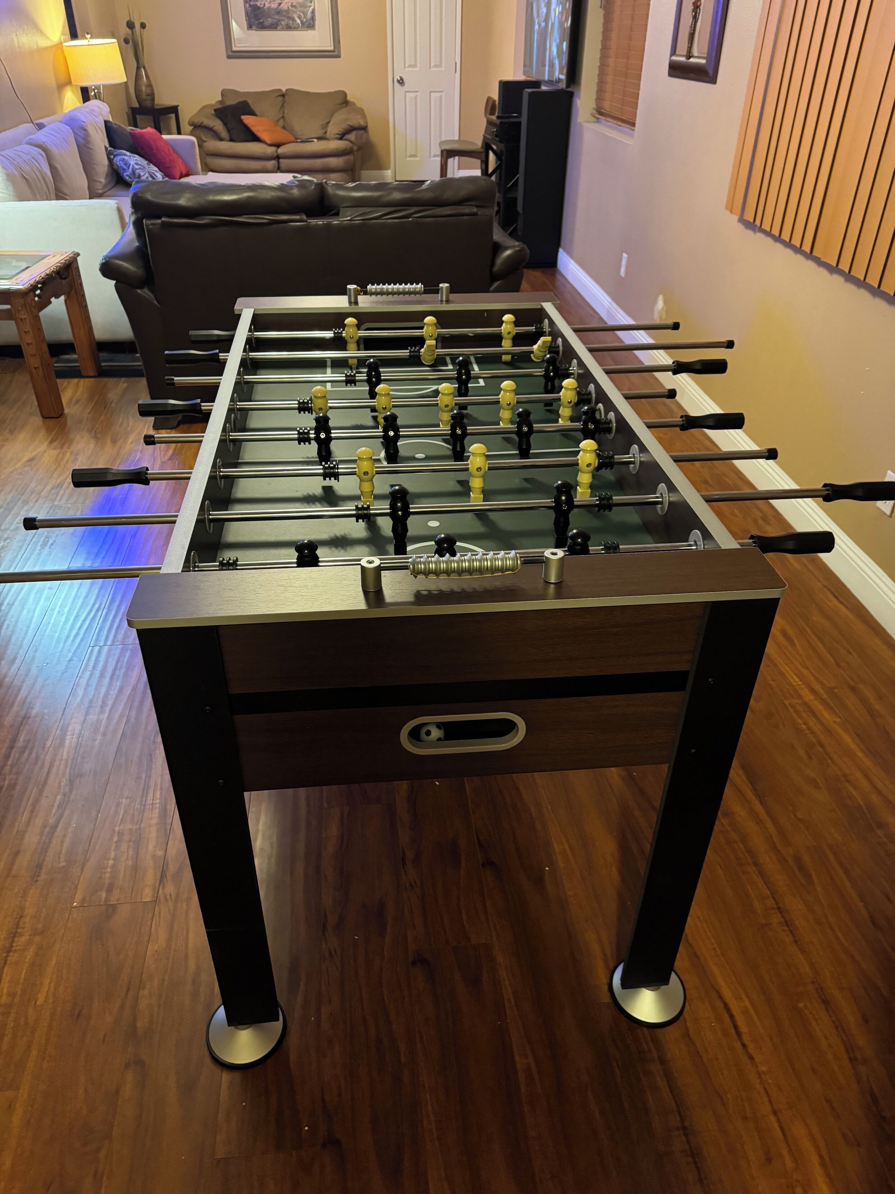 A foosball table in a living room with a sofa and a lamp in the background.