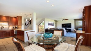 A modern kitchen with wooden cabinets, a dining area with a glass table, and a living room with a TV and seating.