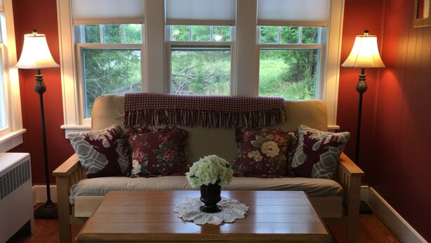 A cozy living room with a sofa, a coffee table, and two lamps. There are three windows with blinds, and a view of greenery outside. - Unique FarmStay Experience in The Catskills! (Durham)