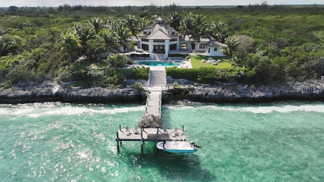 An aerial view of a resort with a dock leading to a house surrounded by lush greenery and a clear turquoise pool.