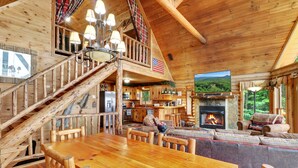 A log cabin interior with a wooden dining table, a fireplace, a kitchen, and a balcony with an American flag. - Luxurious, Secluded Cabin, with Gazebo Covered Hot-tub + Amazing View (Blue Ridge)