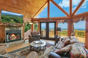 A cozy cabin interior with a stone fireplace, comfortable seating, and a large window offering a scenic mountain view.