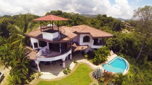 Une grande maison avec une piscine, entourée d’une végétation luxuriante et de palmiers.
