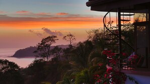 Une vue sur la côte, avec un coucher de soleil, des palmiers et un escalier menant à un balcon orné de fleurs rouges.