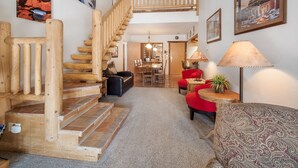 Un escalier en bois menant à une chambre aménagée en mezzanine, un coin repas agrémenté d’un lustre, et un espace salon confortable avec un fauteuil à motifs et un canapé rouge.