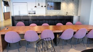 Un comedor moderno con una mesa de madera alargada y sillas moradas, una cocina con armarios blancos e iluminación colgante.