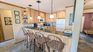 A kitchen with wooden cabinets, a central island with bar stools, pendant lighting, and a refrigerator. - Spacious Condo w/ FREE WiFi, Parking, Heated Pool, Hot Tubs, Skier Shuttles (Avon)