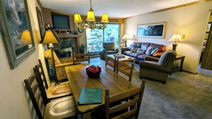 A cozy living room with a fireplace, a dining area with a wooden table and chairs, a leather sofa, and a television mounted on the wall. - Spacious Condo w/ FREE WiFi, Parking, Heated Pool, Hot Tubs, Skier Shuttles (Avon)
