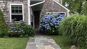 Ein Steinweg führt zu einem Haus mit einer Garagentür, umgeben von üppigem Grün und blühenden Pflanzen.