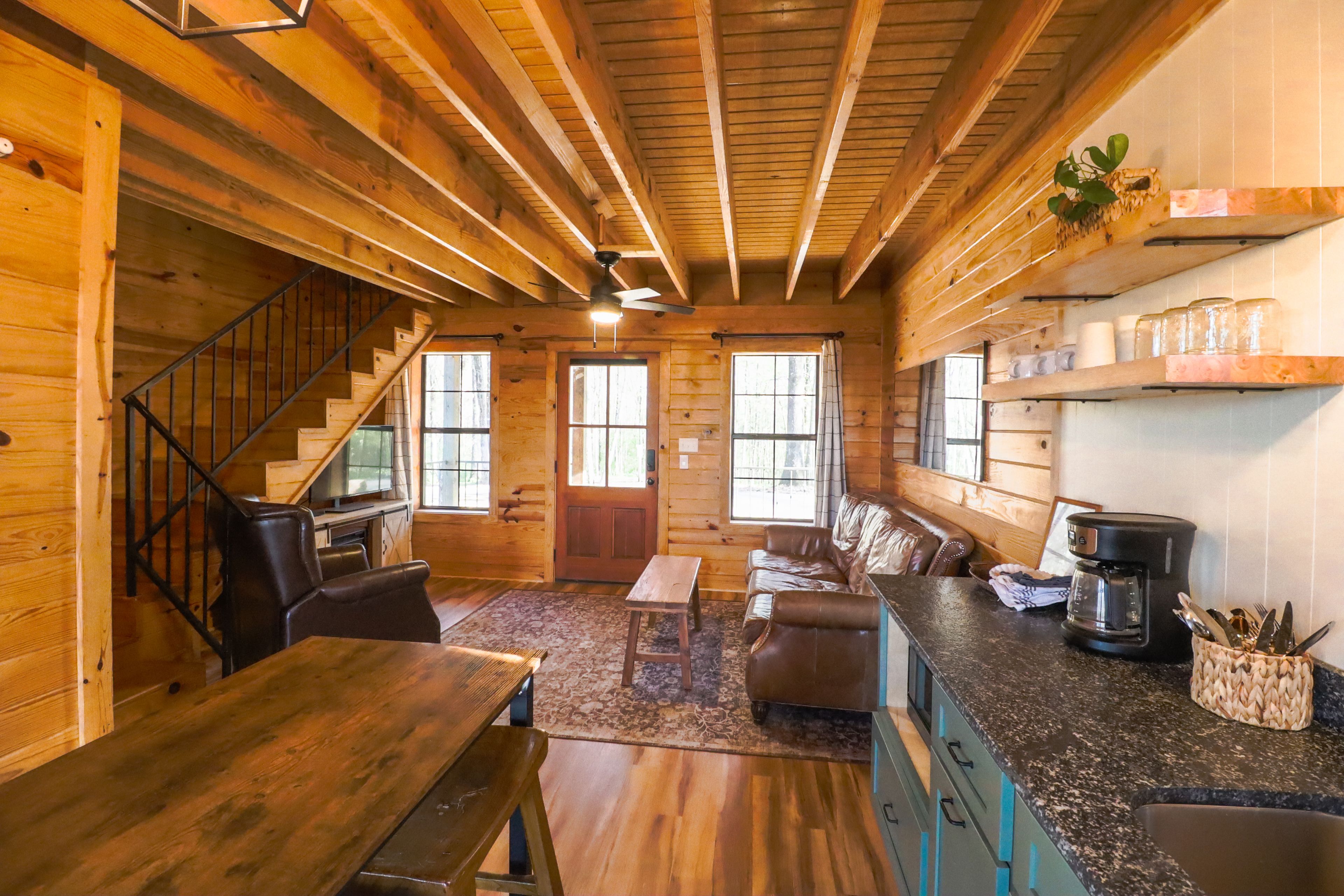 A cozy cabin interior with a wooden staircase, a dining area, a seating area with a sofa, and a kitchen with a coffee maker.