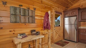 Interior de una cabaña de madera con una mesa de madera, libros, objetos decorativos, un perchero, un refrigerador y una ventana.