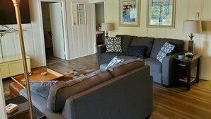 A living room with a sofa, a coffee table, and a lamp. There is a door leading to another room. - Suttons Bay-Pet Friendly-Walk to Brewery, beach, town, shops (Suttons Bay)