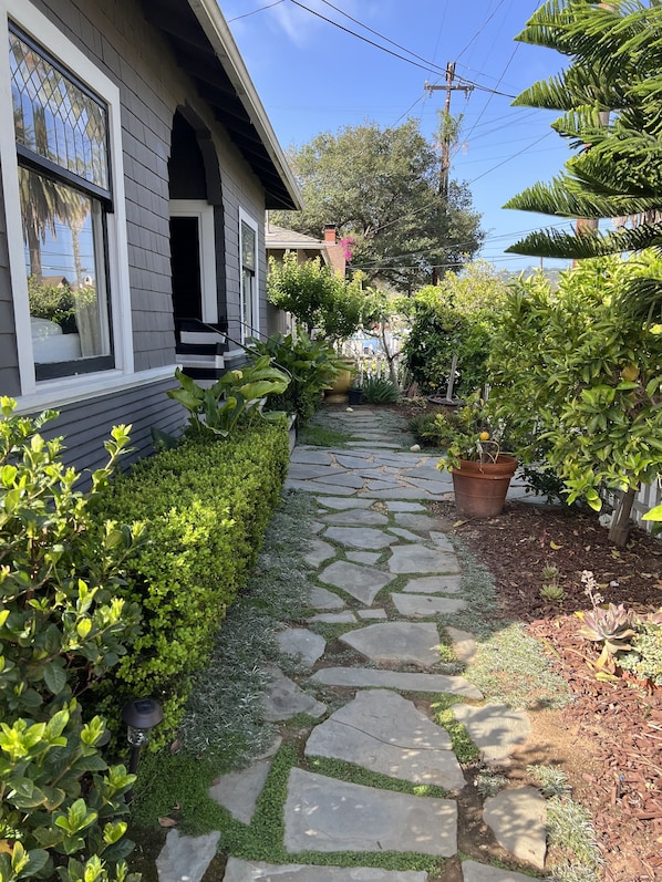 A stone pathway leading to a house with a porch, surrounded by various plants and a small garden area.