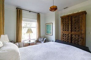 A bedroom with a large bed, a wooden wardrobe, a window with curtains, a framed picture on the wall, and a hanging light fixture.